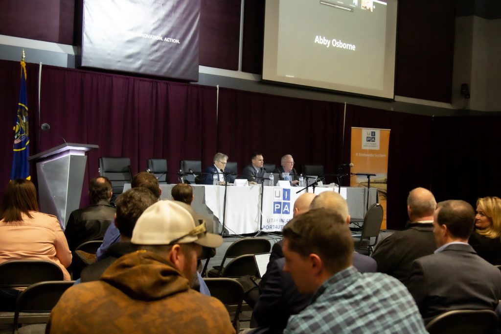 Panel of people sitting on raised platform in front of a maroon curtain, with backs of watching audience in foreground